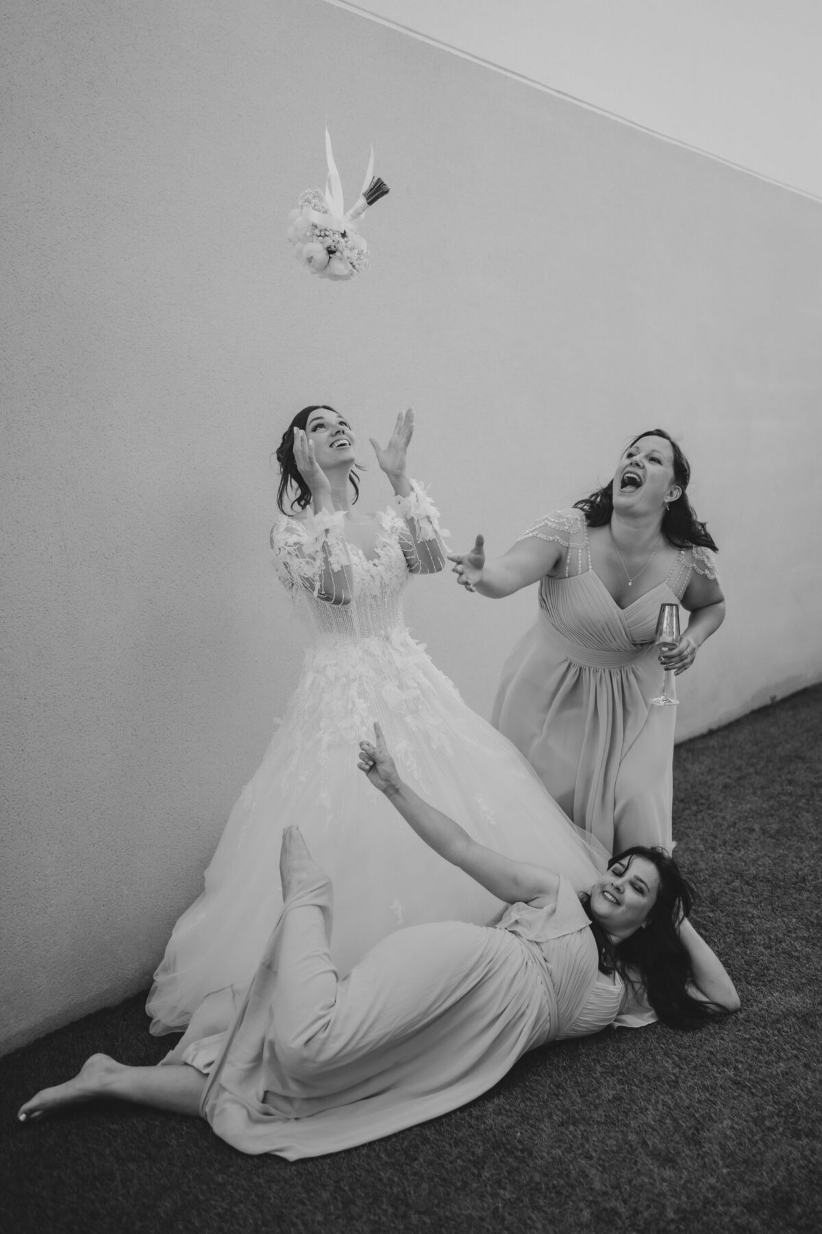 En noir et blanc, la mariée et ses amies rient, assises par terre contre un mur blanc