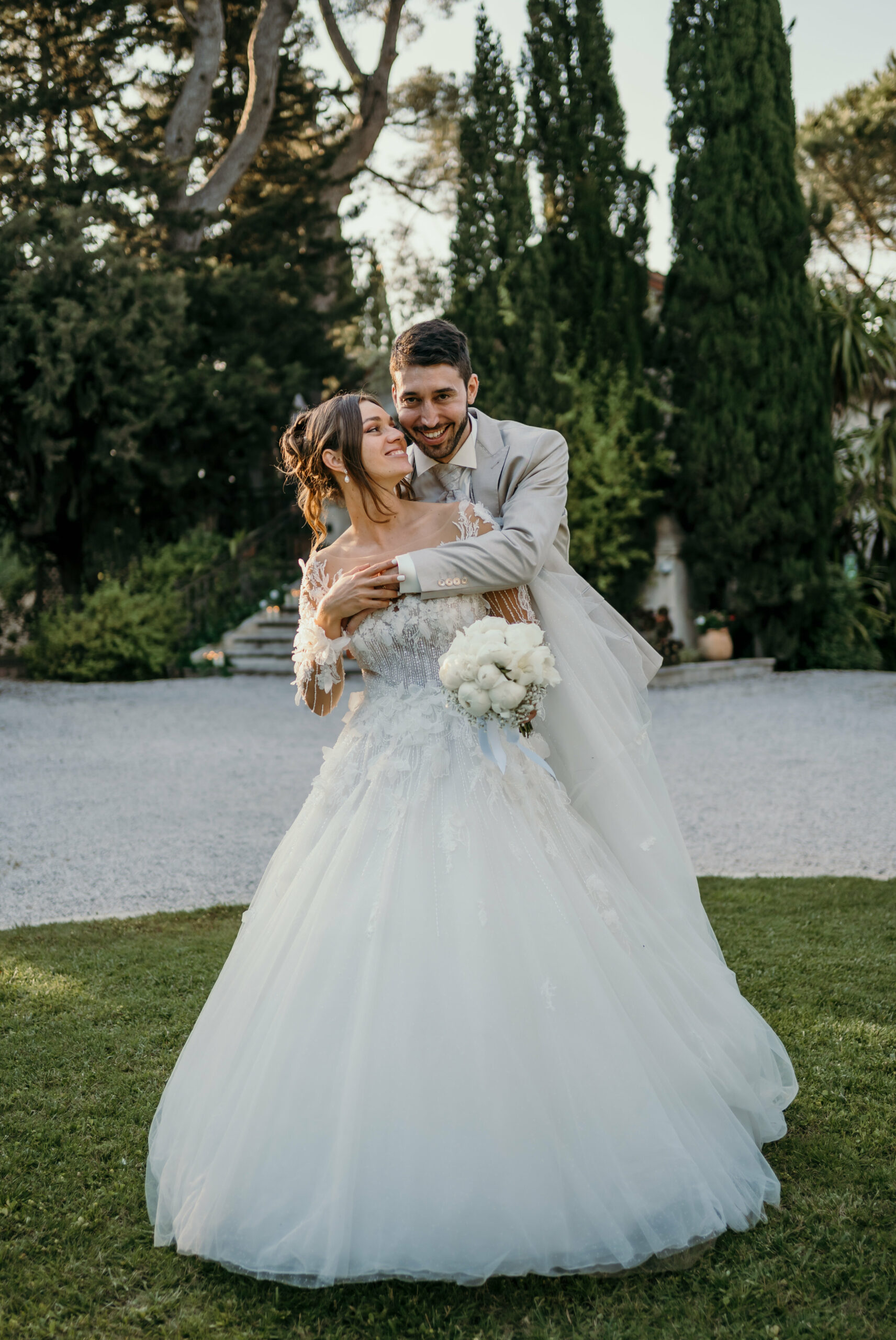 Le marié enlace la mariée en longue robe de tulle, devant une haie de cyprès