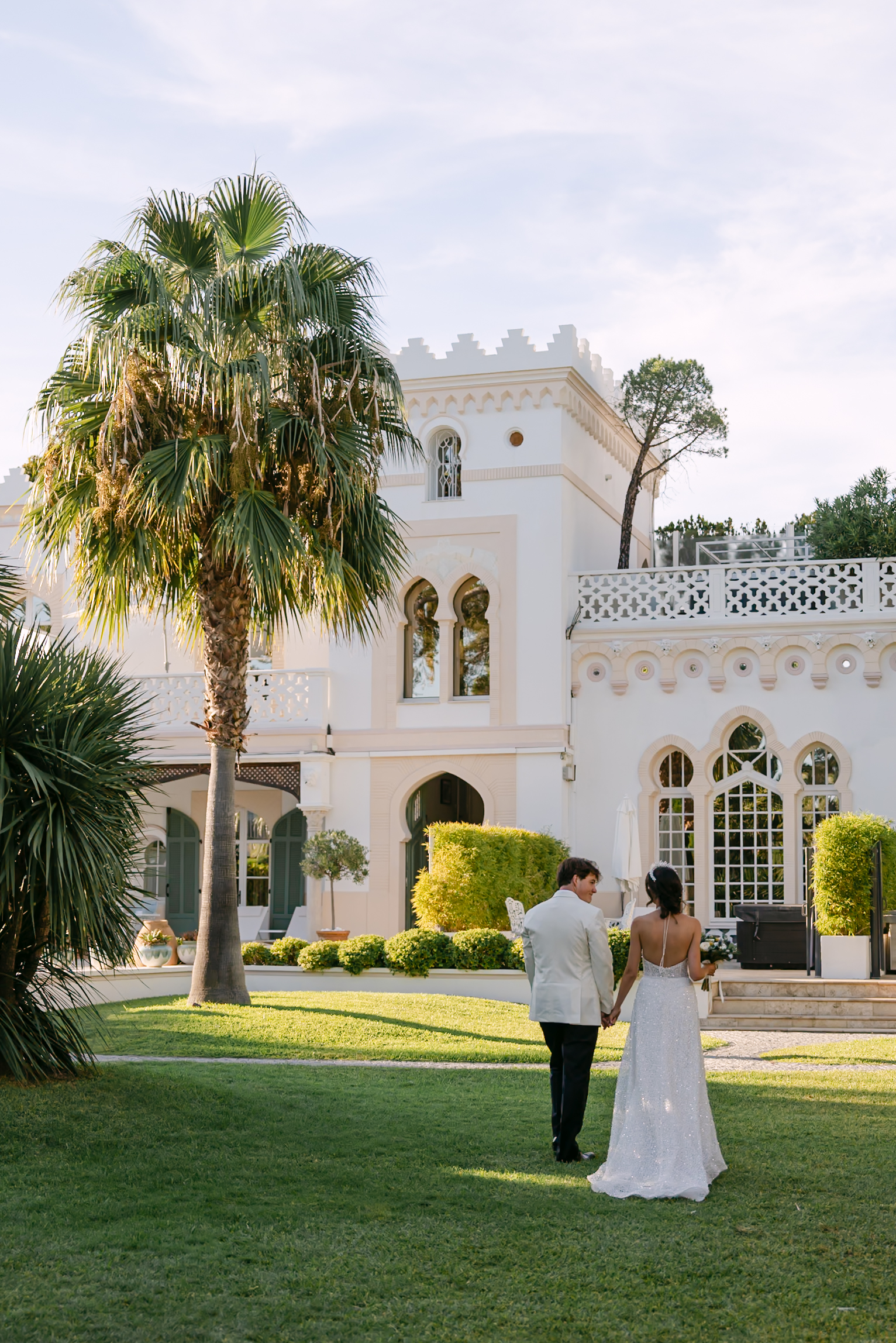 Photographe mariage château Provence 1 Les mariés de dos sur la pelouse d'un château blanc aux créneaux, entre les palmiers