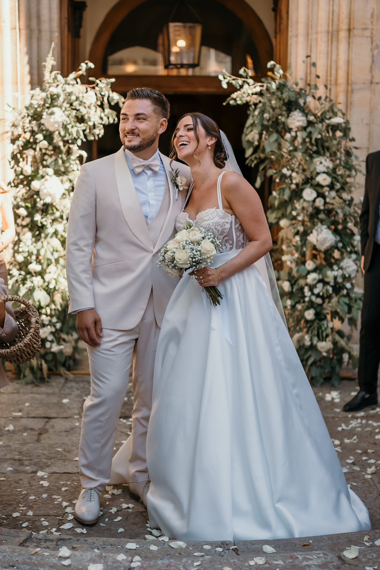 Photographe mariage château Provence 8 Les mariés sortent sous une arche de fleurs blanches, le marié en costume beige