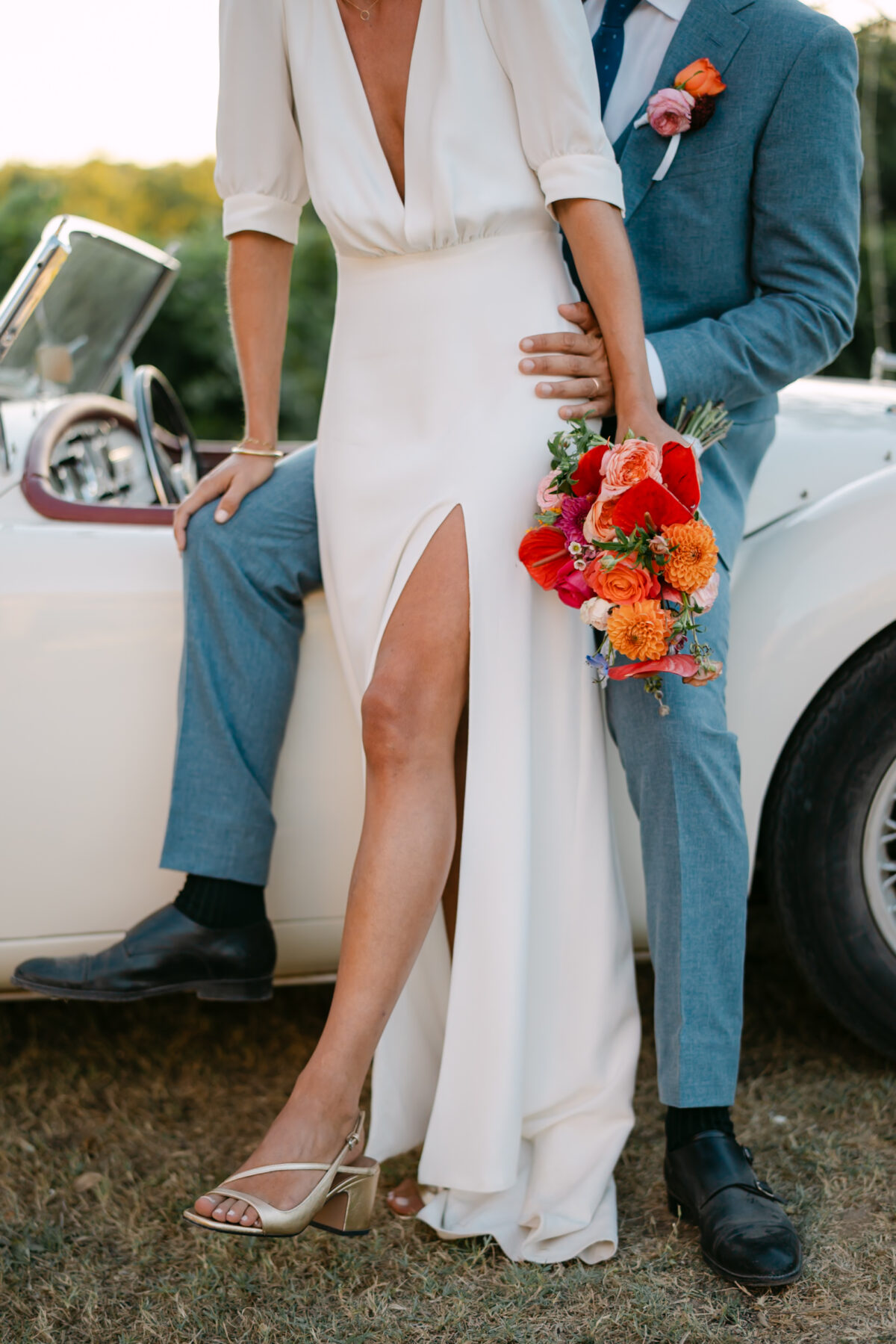 La mariée appuyée contre une voiture ancienne, un bouquet orange contre le costume bleu du marié