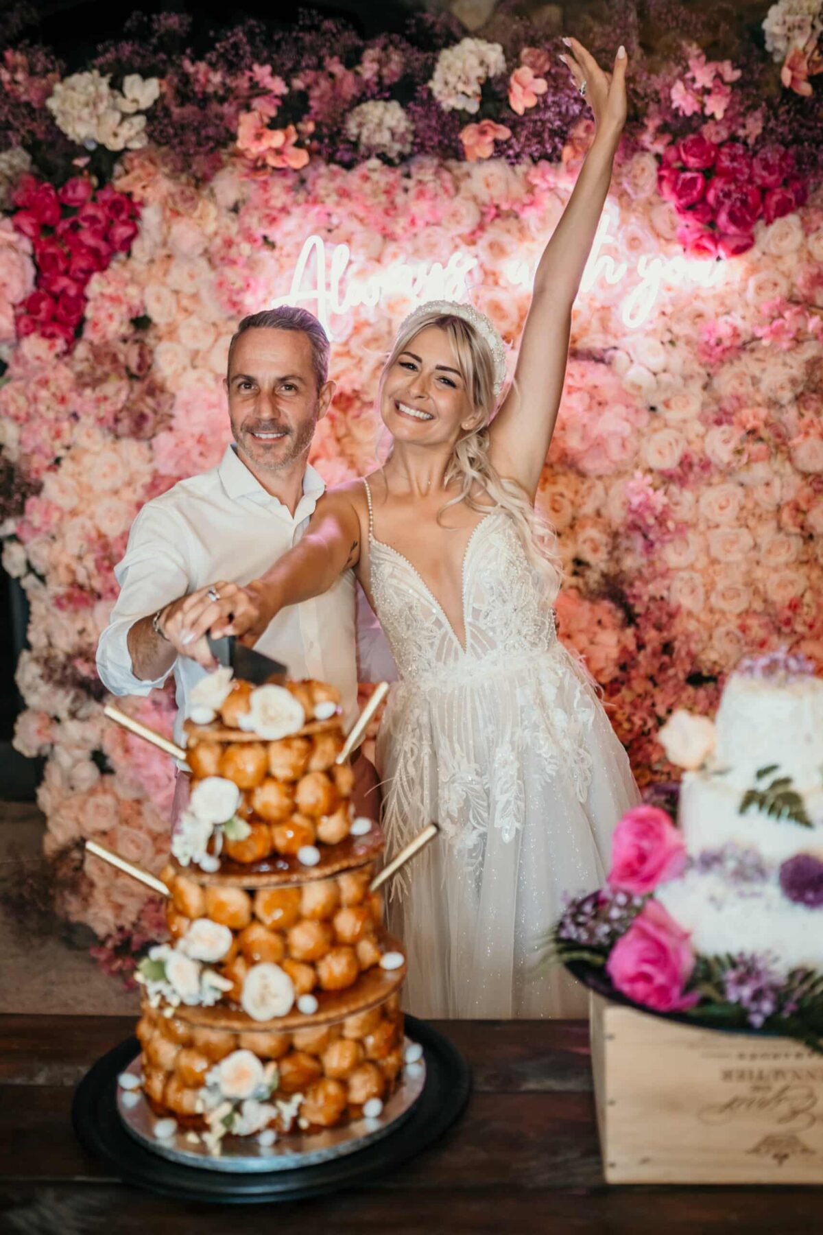 Les mariés lèvent les bras devant un mur de fleurs roses, à côté de la pièce montée