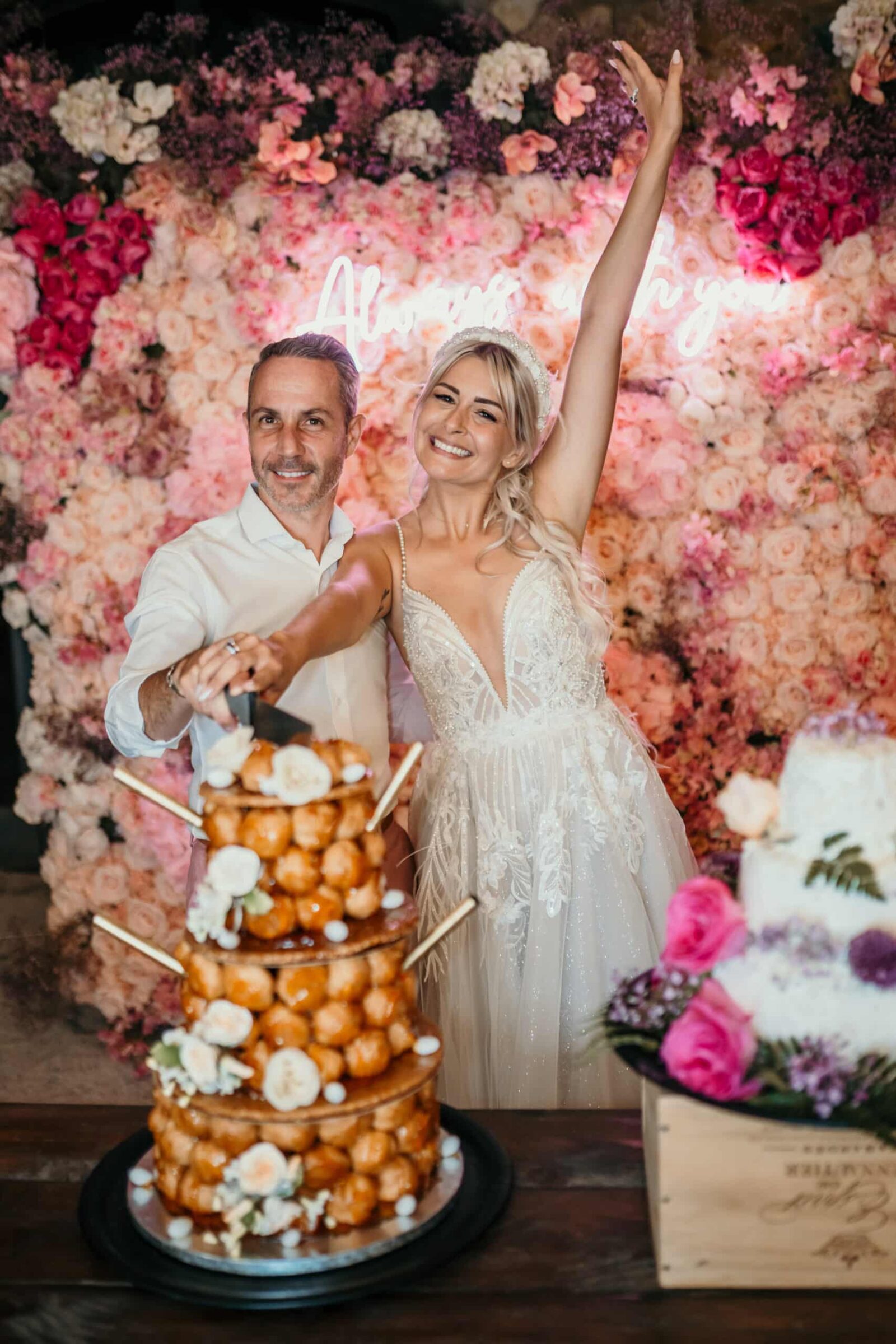 Photographe mariage Les Arcs 9 Les mariés lèvent les bras devant un mur de fleurs roses, à côté de la pièce montée