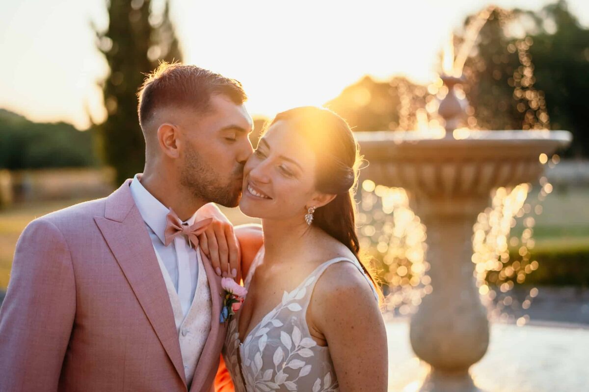 Le marié en costume rose embrasse la mariée devant une fontaine, au coucher du soleil