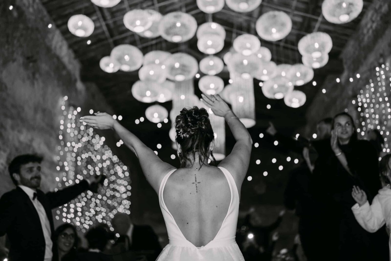 Photographe mariage Les Arcs 10 En noir et blanc, la mariée de dos danse sous des suspensions lumineuses