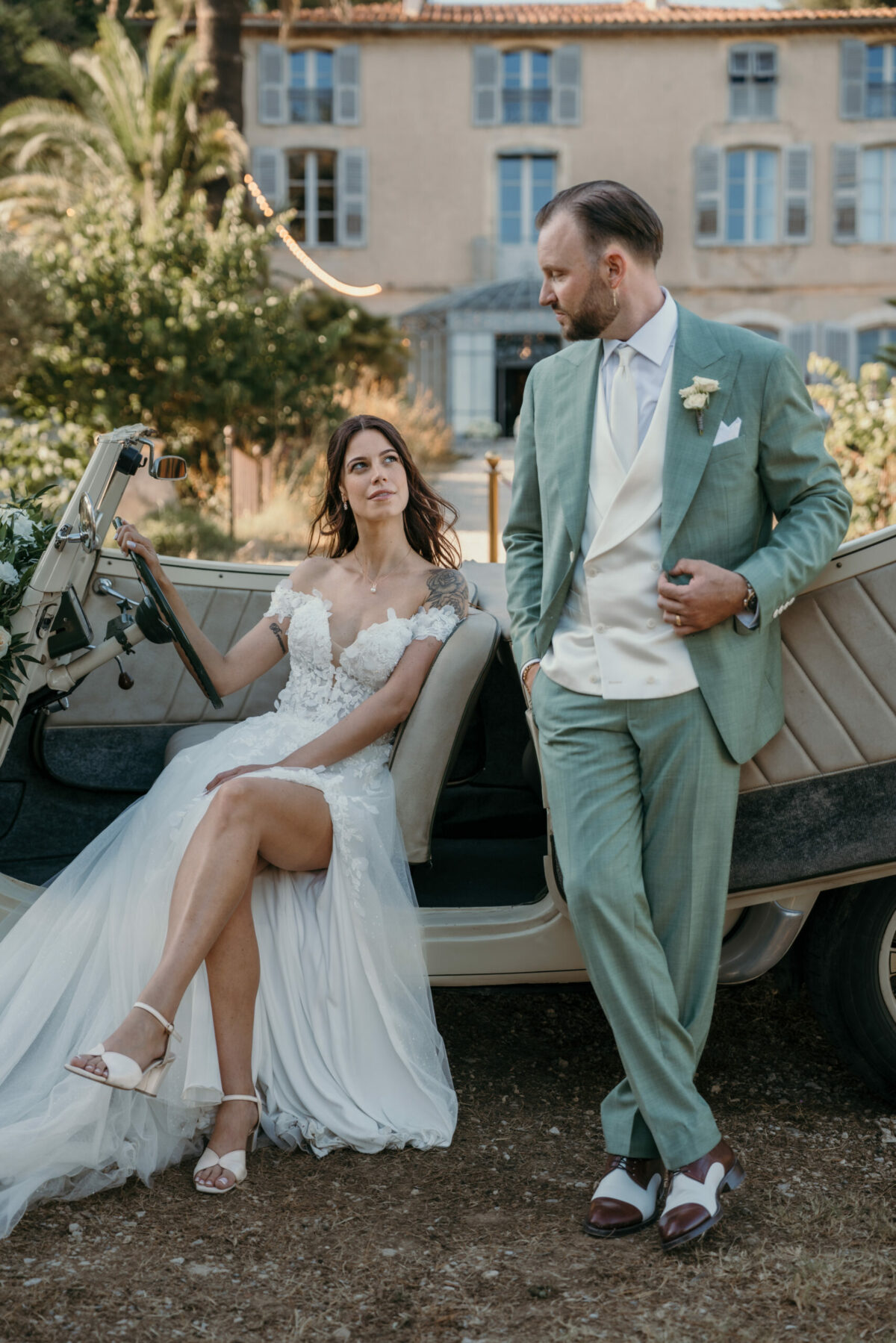 La mariée assise sur une voiture ancienne, le marié en costume vert d'eau debout à côté, devant une bastide