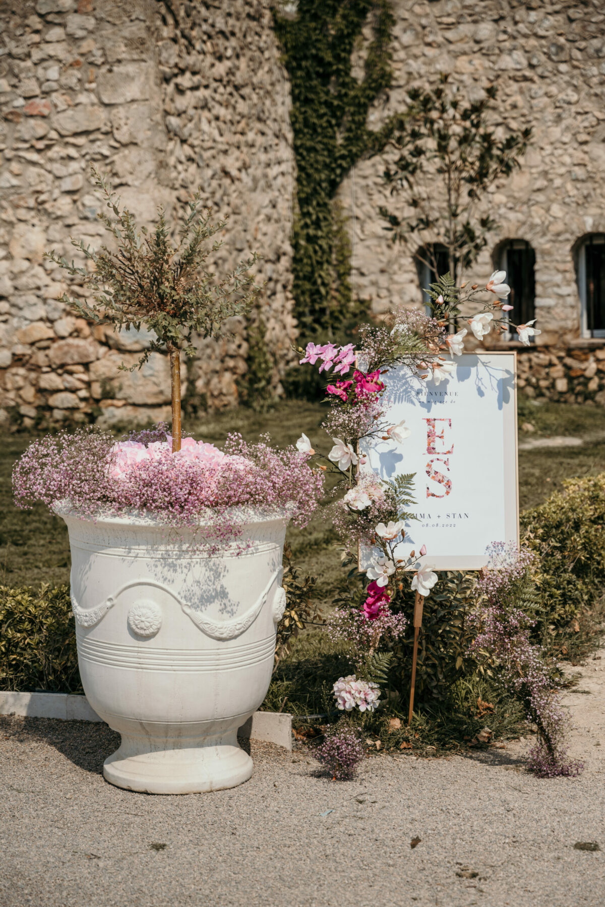Un vase d'Anduze fleuri et le panneau d'accueil, devant un mur de pierre ancien