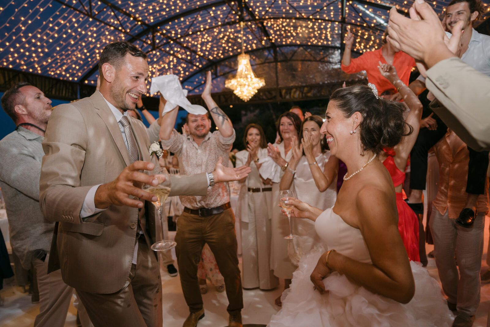 Accueil 19 Le marié et la mariée dansent au milieu des invités qui lèvent les bras, sous un lustre et une voûte de lumières