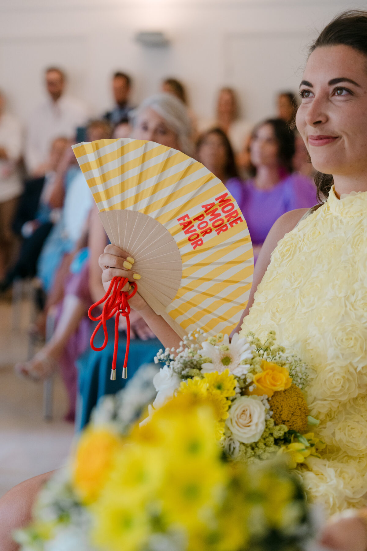 Accueil 4 Une invitée sourit derrière un éventail jaune et rose, un bouquet de fleurs jaunes au premier plan