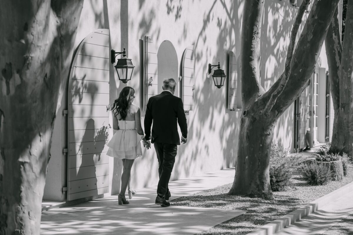 En noir et blanc, la mariée marche au bras d'un proche, sous les platanes, le long de la bastide