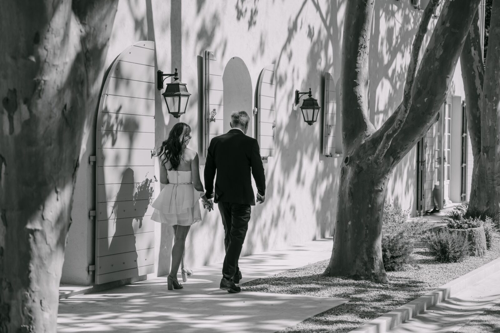 Photographe mariage Salernes 6 En noir et blanc, la mariée marche au bras d'un proche, sous les platanes, le long de la bastide