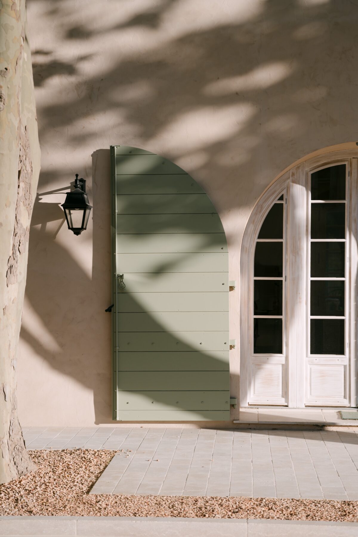 Façade claire d'une bastide, volets vert pâle et lanterne, dans l'ombre des platanes
