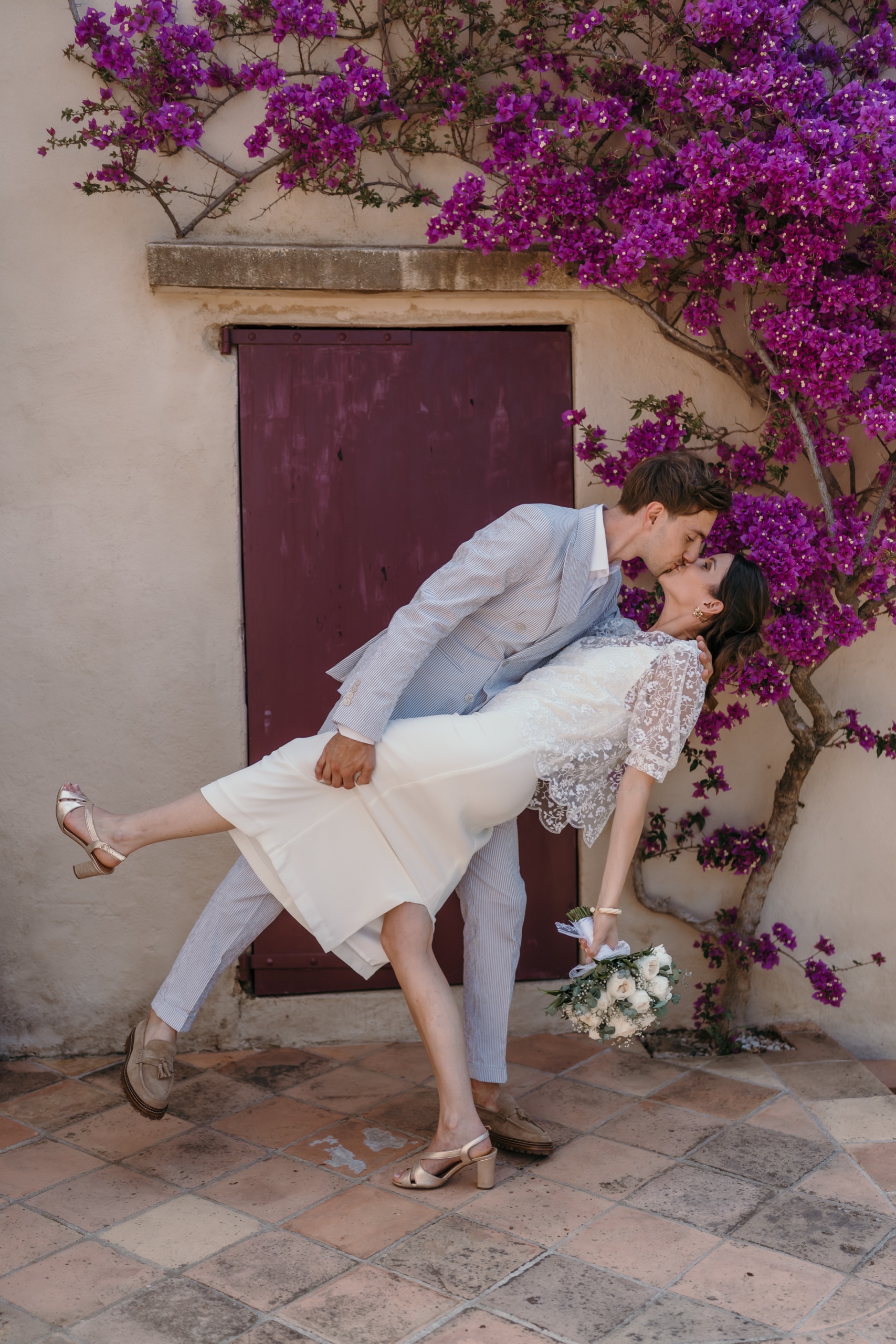 Devant une porte rouge et un bougainvillier en fleurs, le marié fait basculer la mariée pour l'embrasser