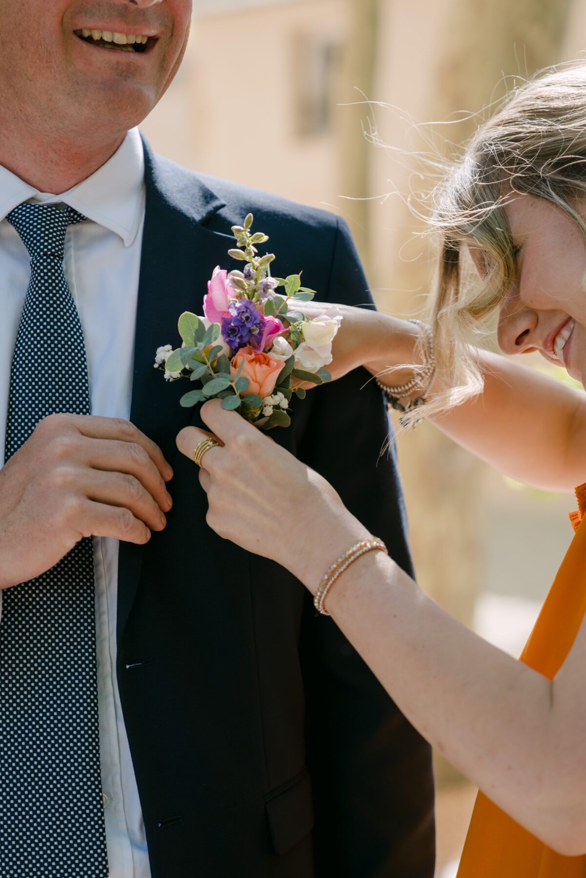 Une invitée en robe orange accroche une boutonnière fleurie au revers d'un invité qui sourit