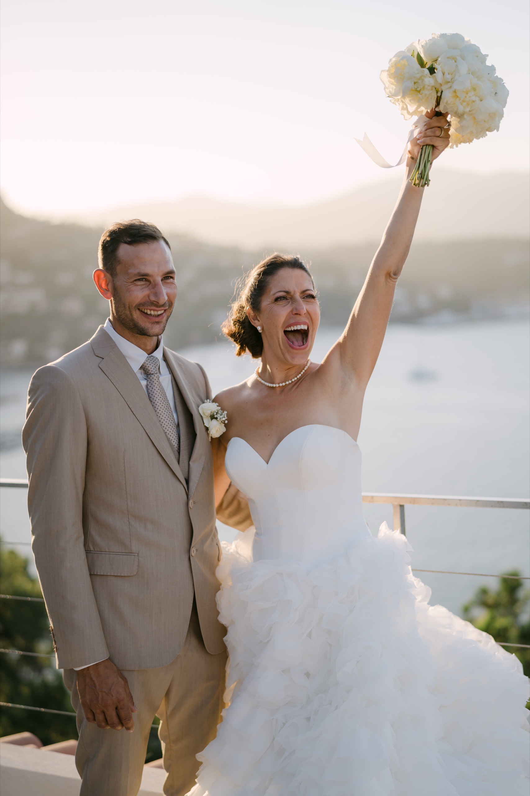 Photographe mariage Saint-Raphaël 1 La mariée lève son bouquet en riant à côté du marié, la mer et le soleil couchant derrière eux