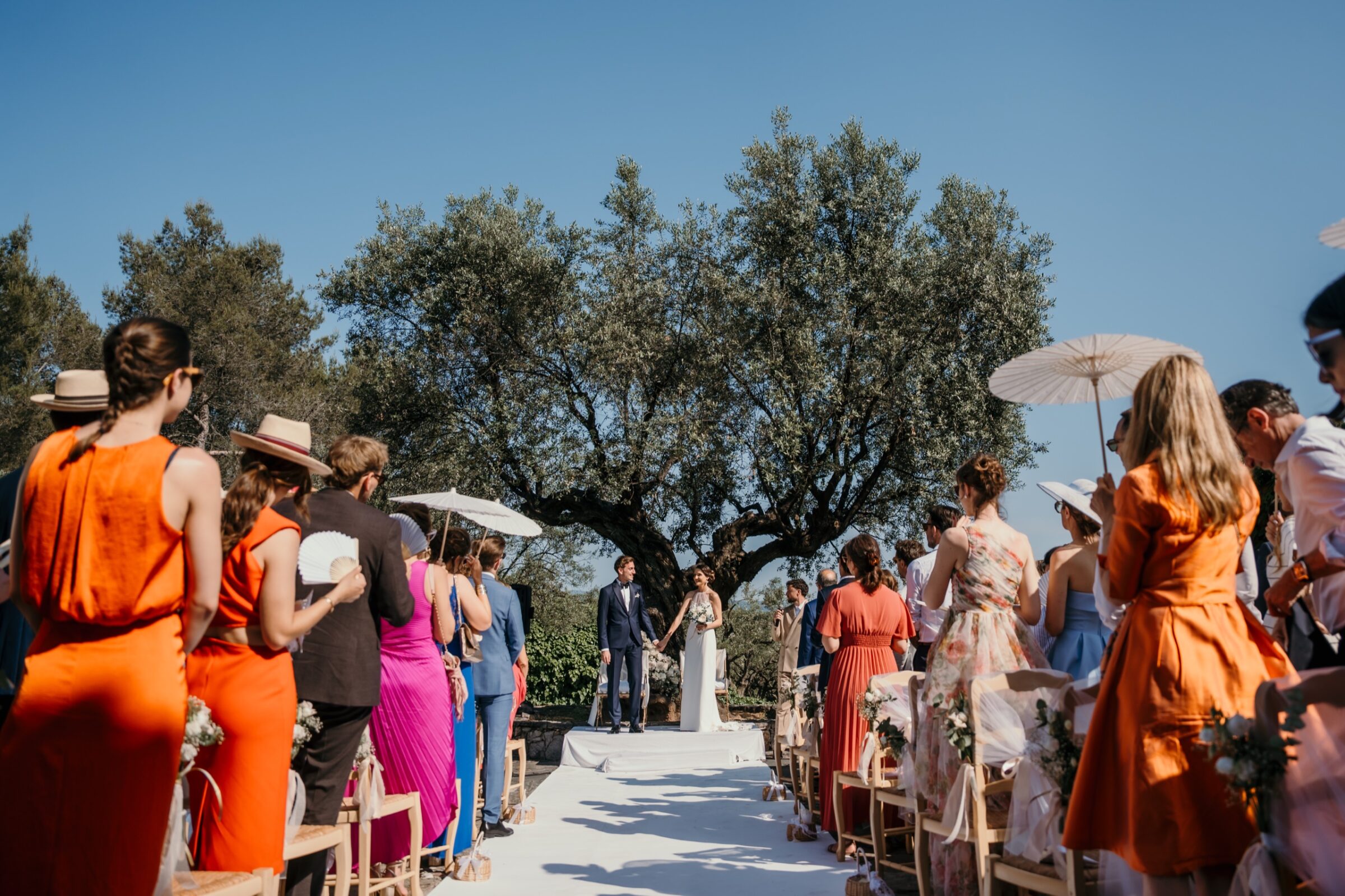 Mon approche 1 Cérémonie en plein air sous les oliviers, invités en robes orange, roses et bleues le long de l'allée