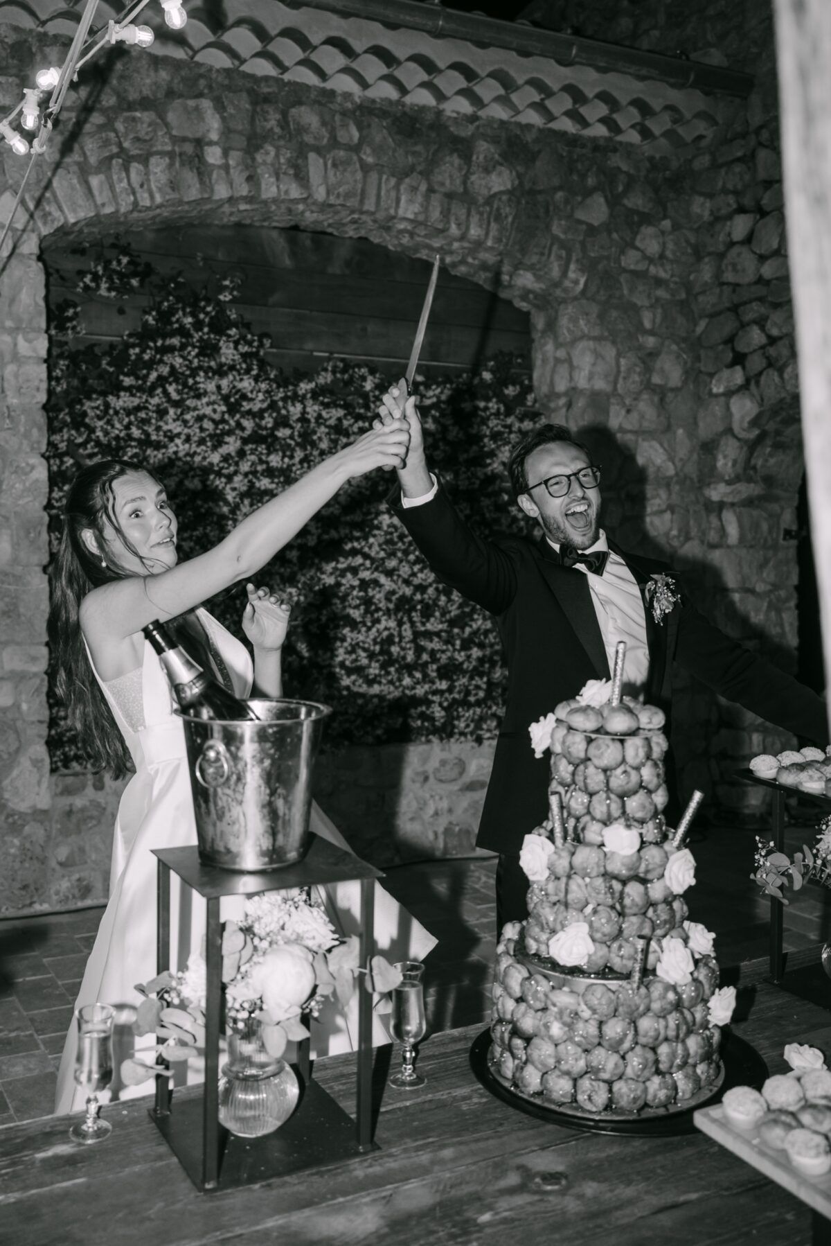Après le mariage : tri, retouche et livraison de vos photos 2 En noir et blanc, les mariés sabrent le champagne devant la pièce montée, dans une salle en pierre