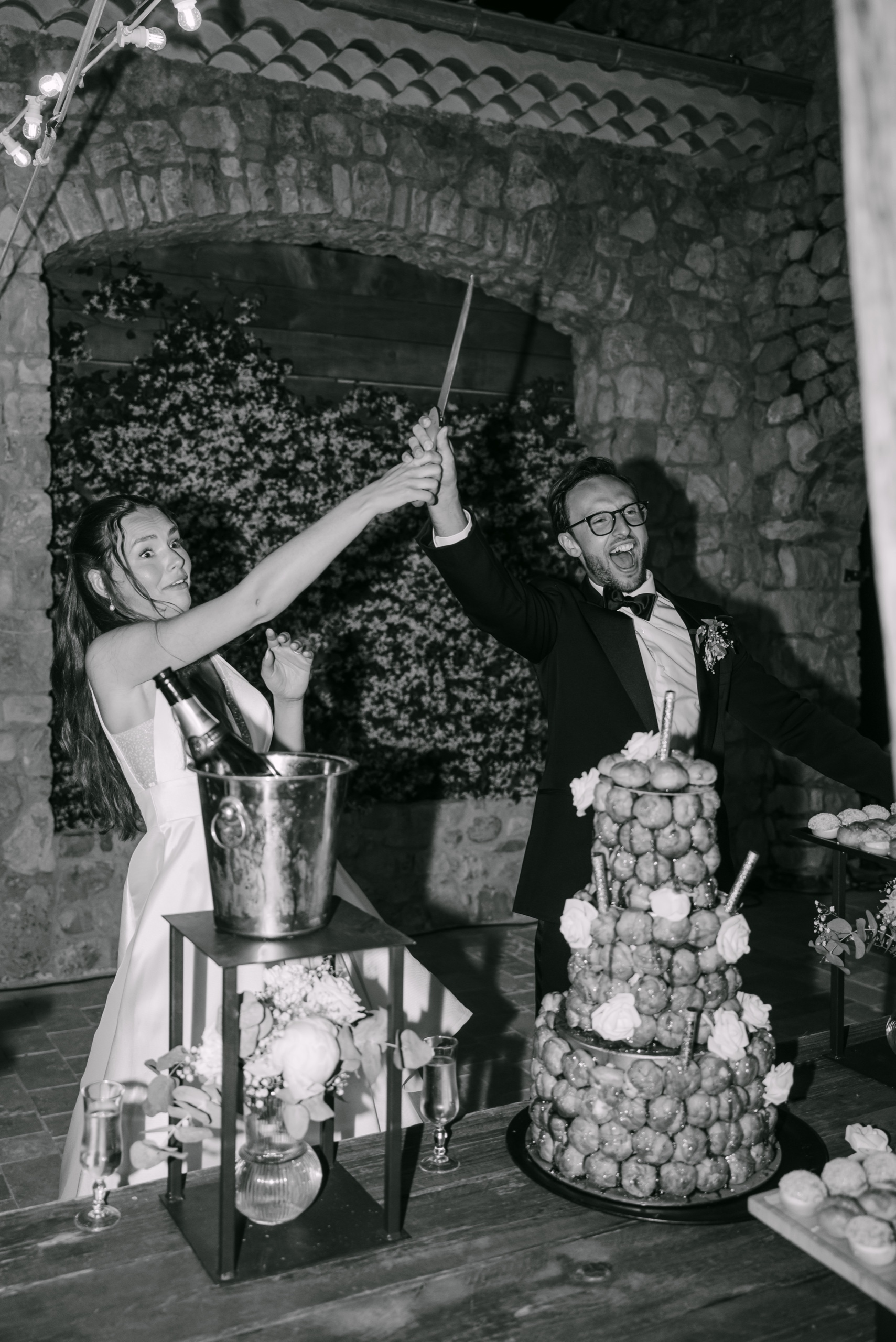 Après le mariage : tri, retouche et livraison de vos photos 4 En noir et blanc, les mariés sabrent le champagne devant la pièce montée, dans une salle en pierre