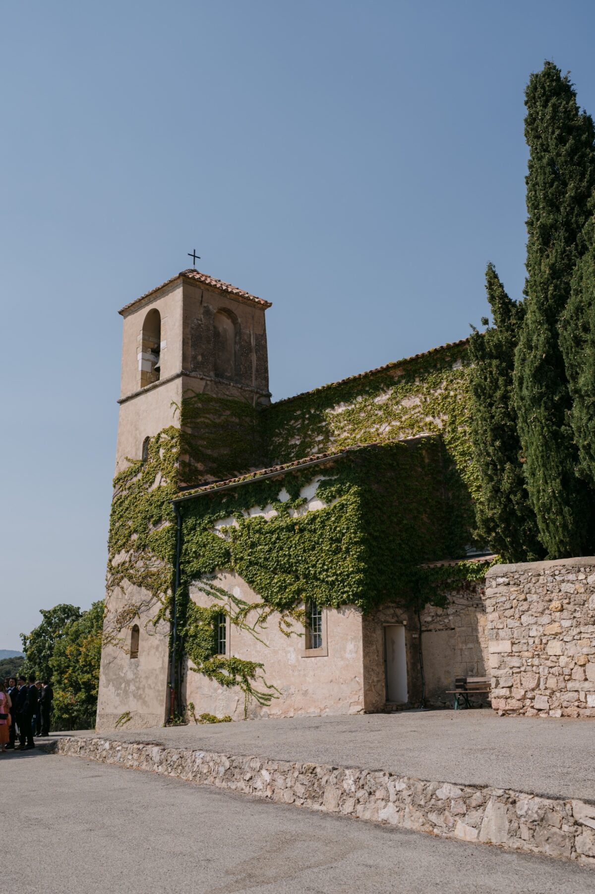 Une chapelle en pierre couverte de lierre, son clocher et deux cyprès sous le ciel bleu