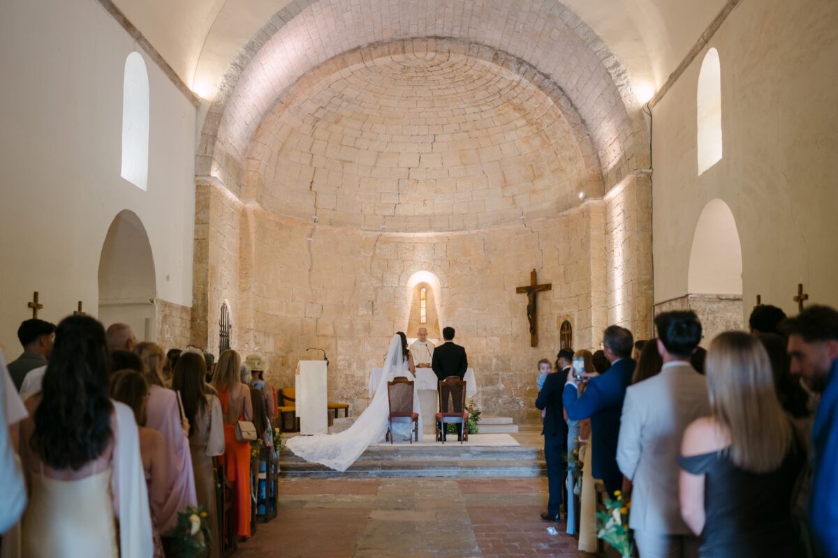 Cérémonie dans une chapelle romane aux murs de pierre claire, les invités debout de part et d'autre de l'allée