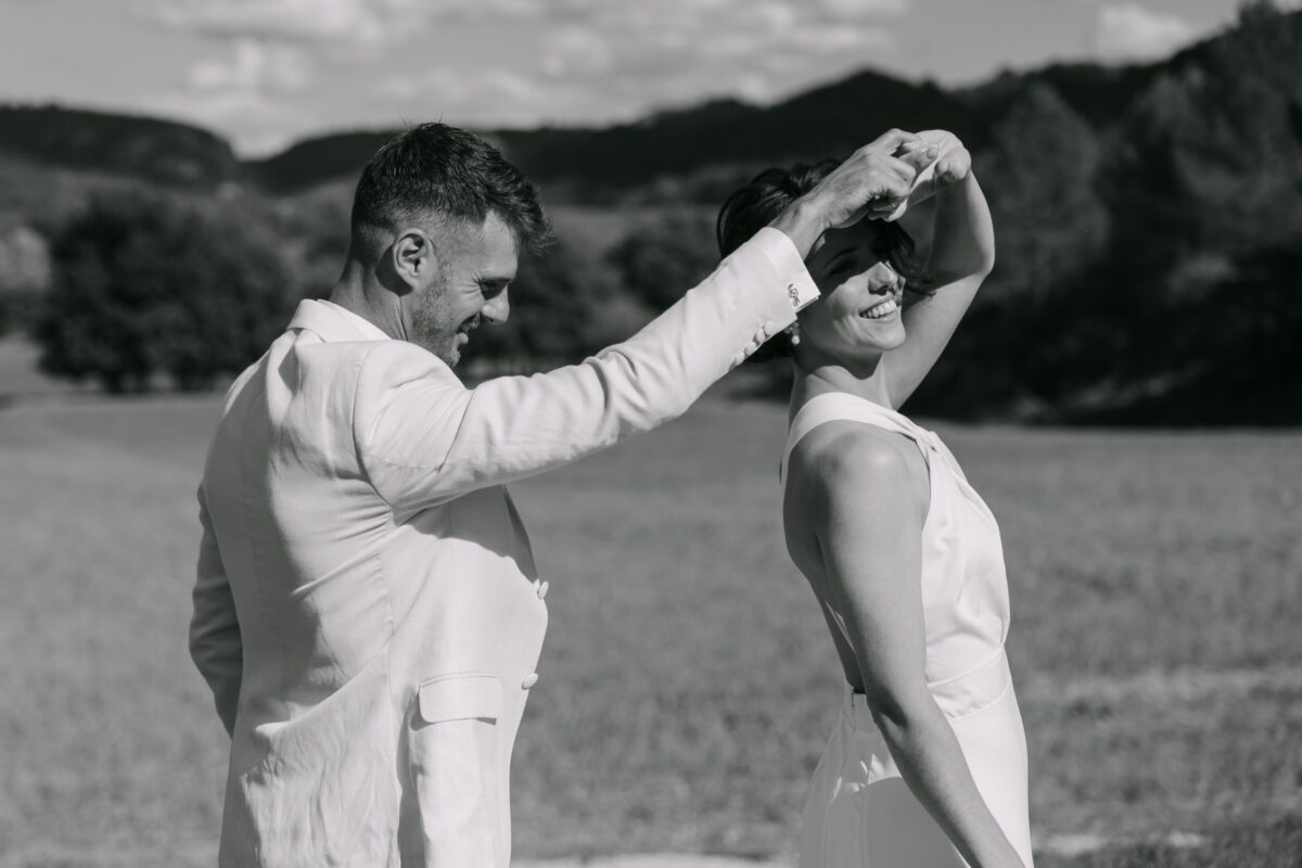 En noir et blanc, le marié fait tourner la mariée au milieu d'un champ, les collines à l'horizon