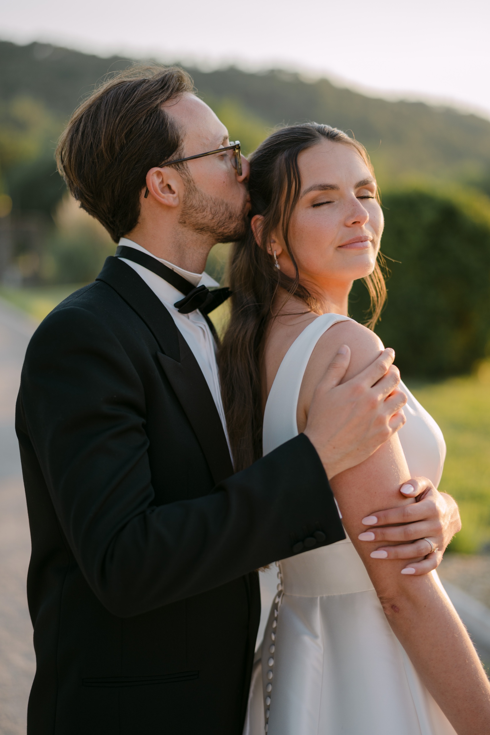 Le marié embrasse la mariée sur la tempe, elle ferme les yeux, les collines derrière eux