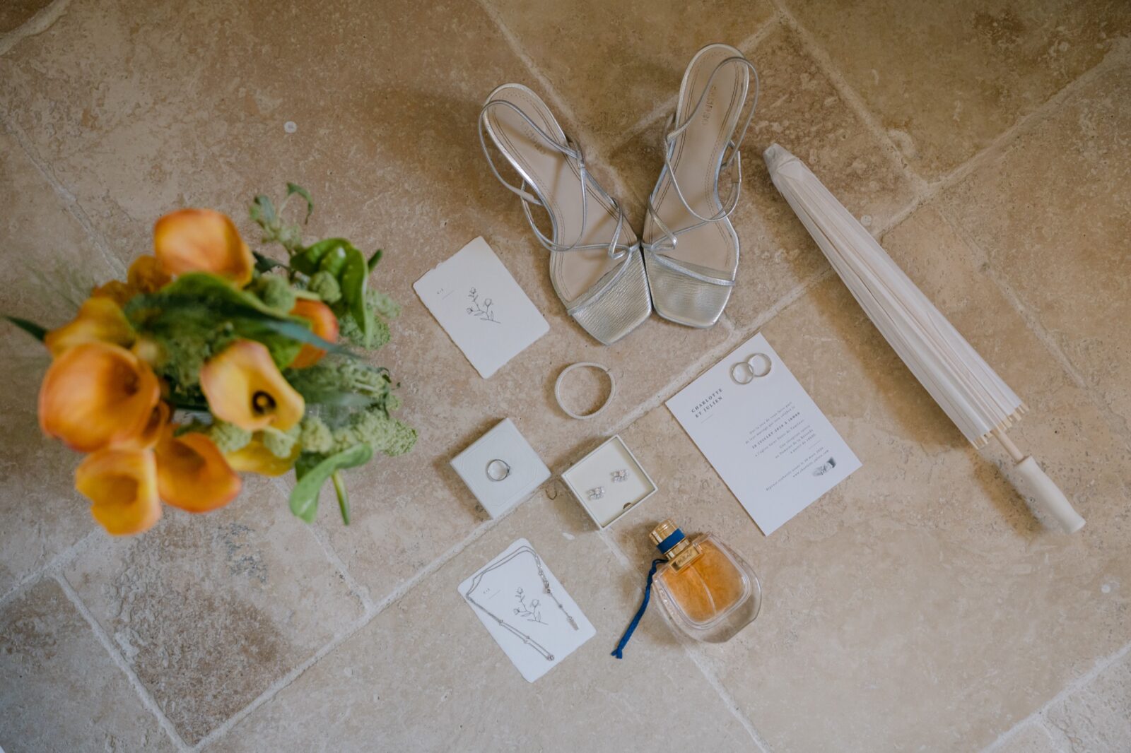Photographe mariage Cogolin 4 Bouquet de callas orange, chaussures, faire-part et parfum posés sur une pierre claire