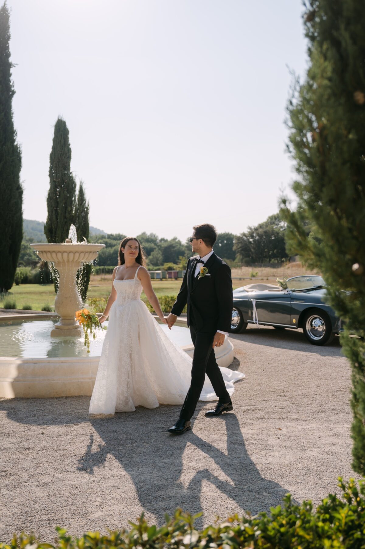 Comment choisir son photographe de mariage : 7 questions à lui poser 5 Les mariés marchent vers une fontaine entre les cyprès, la Porsche ancienne garée à côté