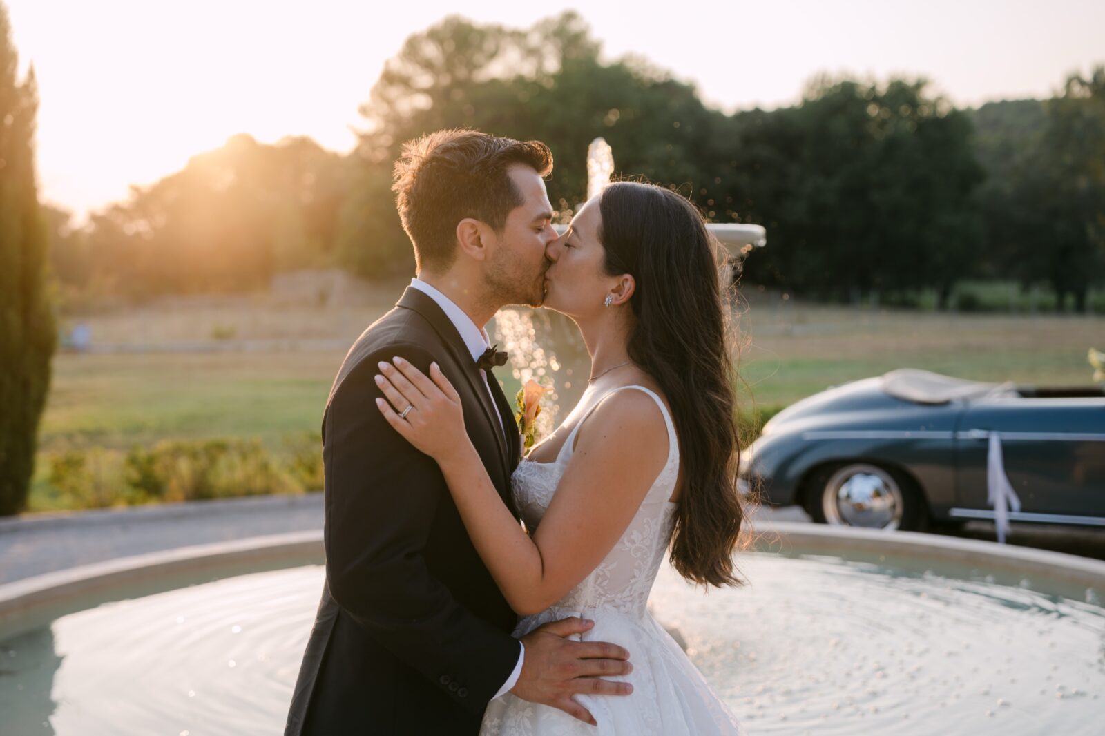 Photographe mariage haut de gamme Provence 3 Les mariés s'embrassent devant une fontaine au coucher du soleil, une Porsche ancienne garée derrière eux
