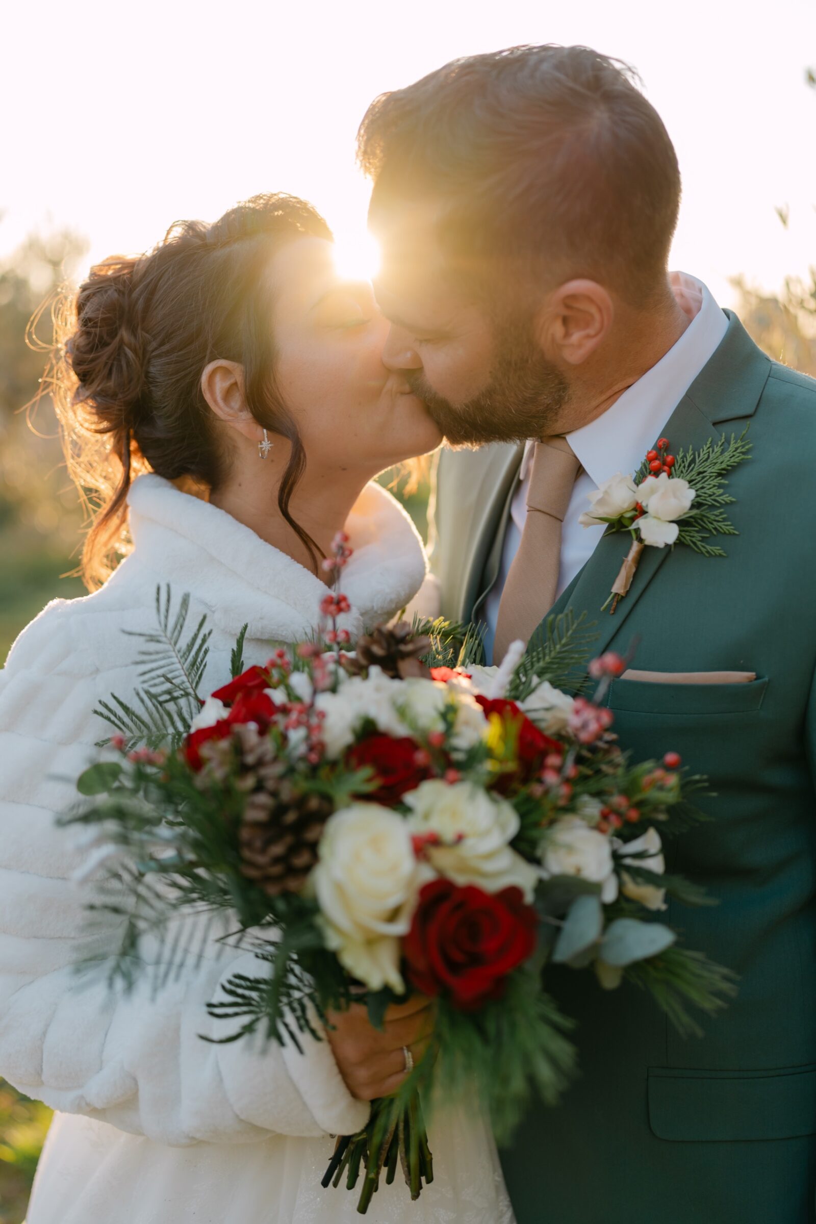 Accueil 11 Mariage d'hiver : les mariés s'embrassent à contre-jour, un bouquet de roses rouges et de branches de sapin entre eux