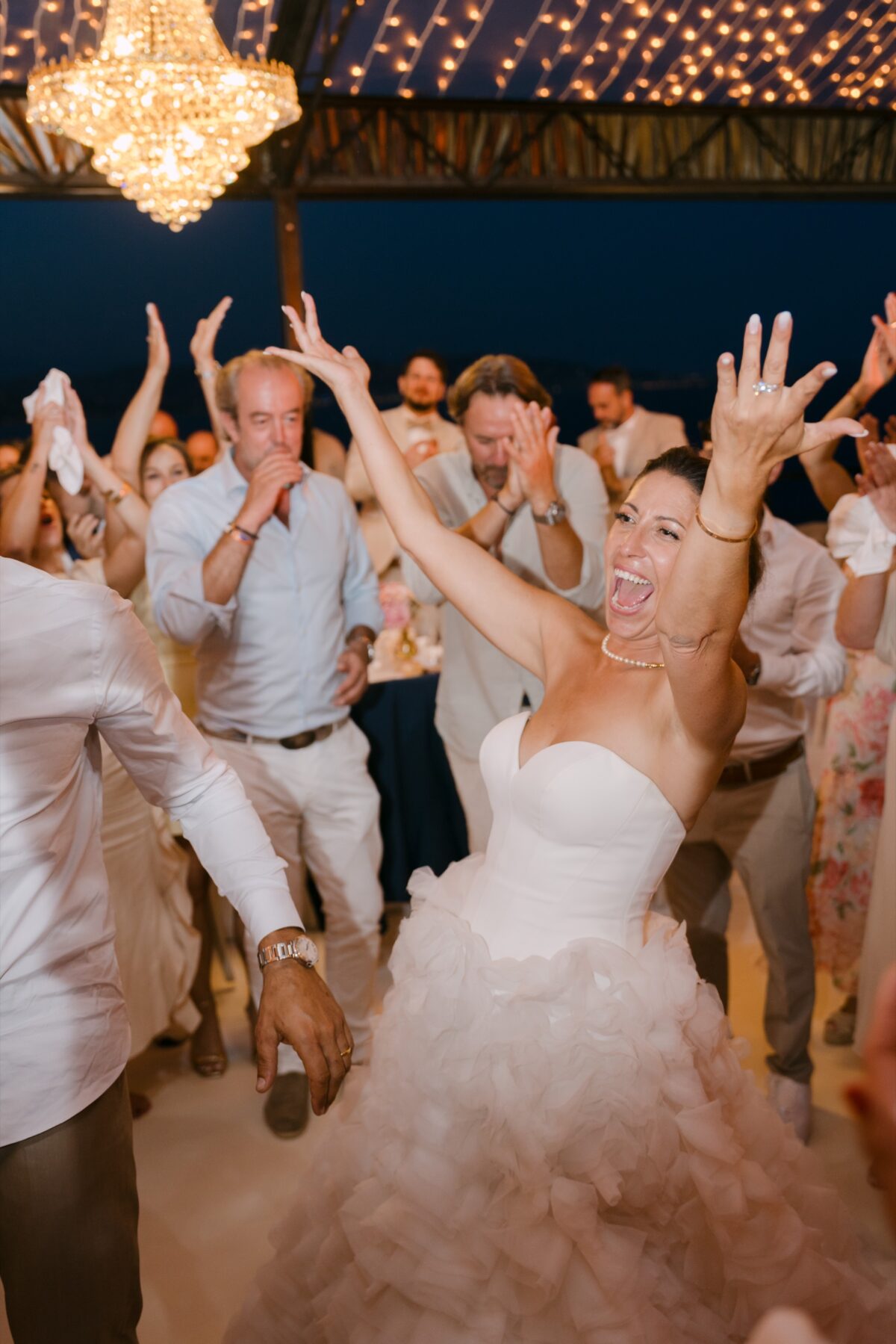 La mariée danse les bras levés en riant, entourée des invités, sous un lustre en cristal