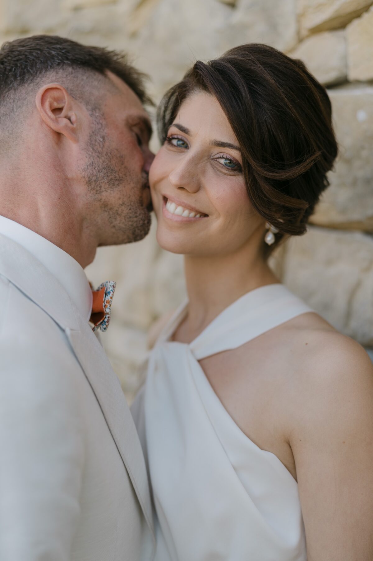 Le marié embrasse la mariée sur la joue, elle sourit, devant un mur de pierre