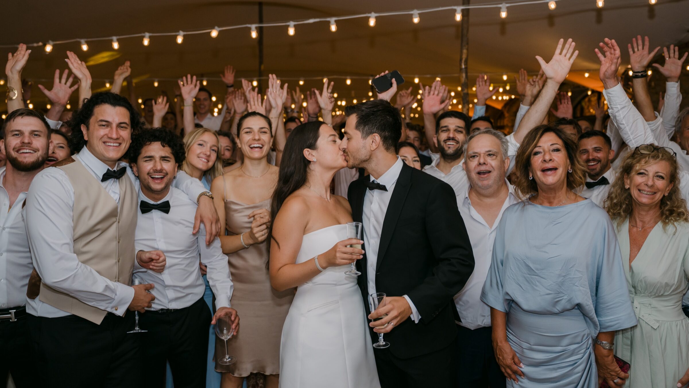 Combien de temps prévoir son photographe le jour du mariage ? 4 Les invités lèvent les bras autour des mariés qui s'embrassent sous les guirlandes
