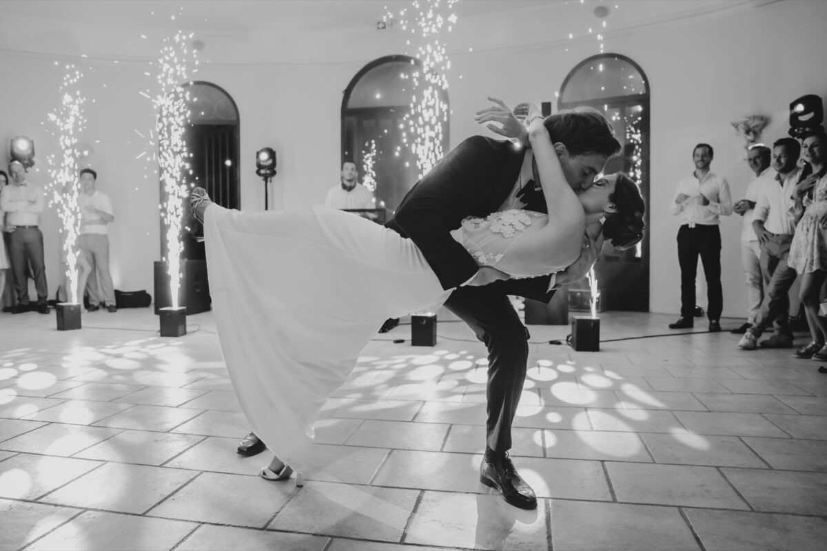 En noir et blanc, le marié fait basculer la mariée pendant la première danse, entouré des invités