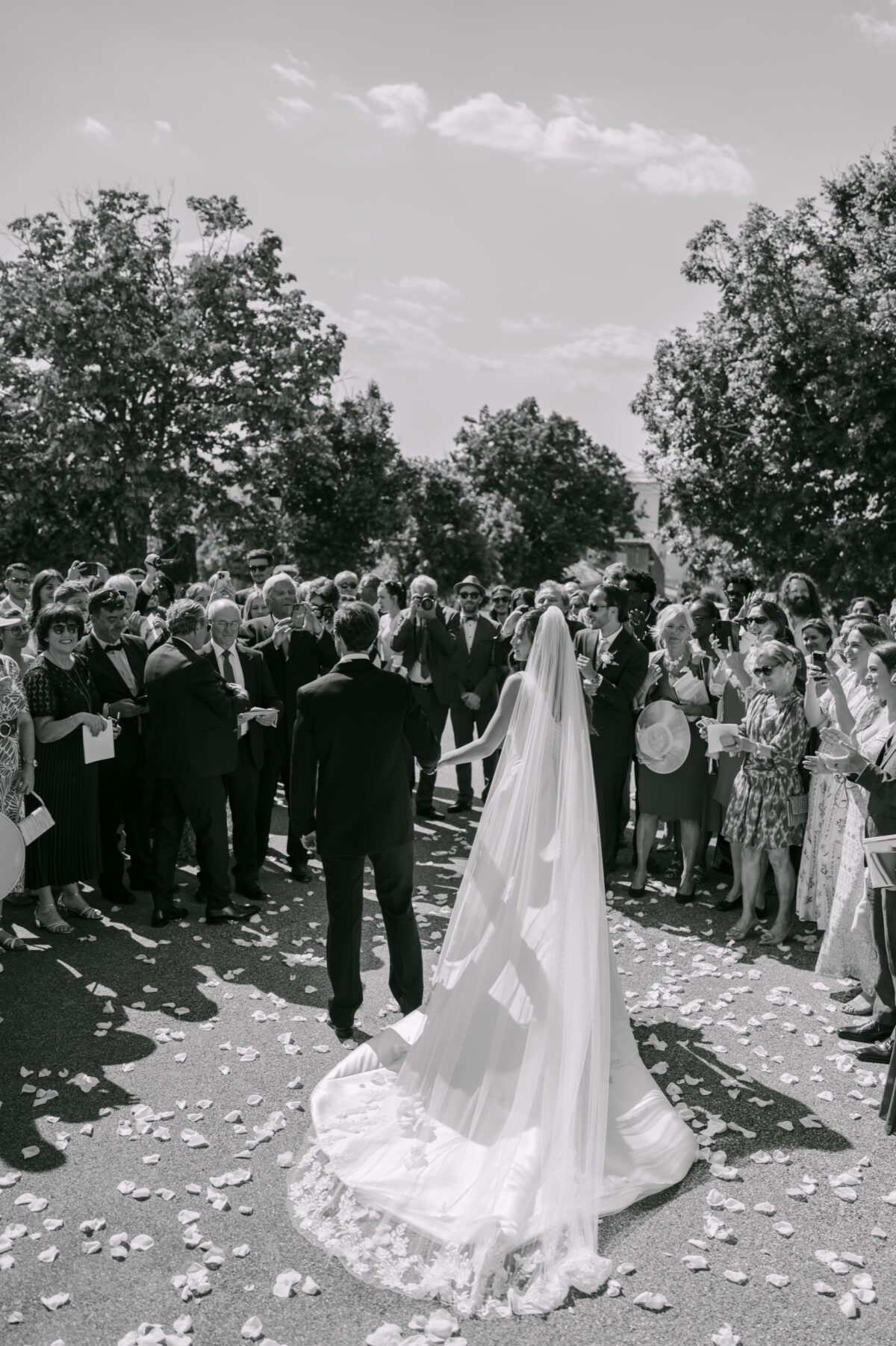 En noir et blanc, les mariés sortent de l'église au milieu des invités, la longue traîne sur les pétales