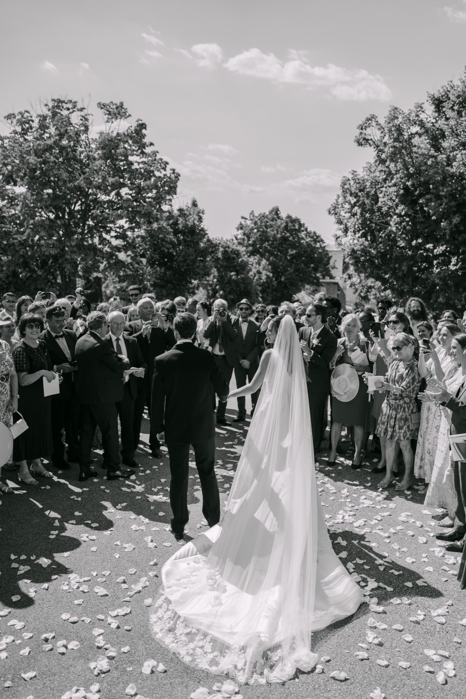 Photographe mariage Fréjus 3 En noir et blanc, les mariés sortent de l'église au milieu des invités, la longue traîne sur les pétales