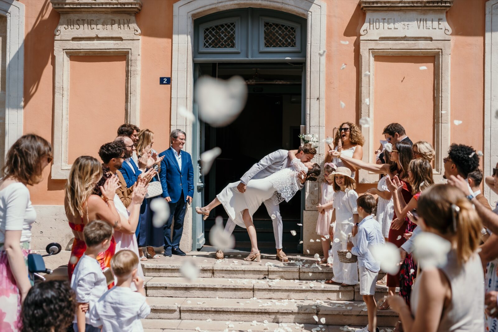 Photographe mariage Draguignan 3 Sur les marches d'une mairie aux murs ocre, le marié fait basculer la mariée sous les pétales lancés par les invités