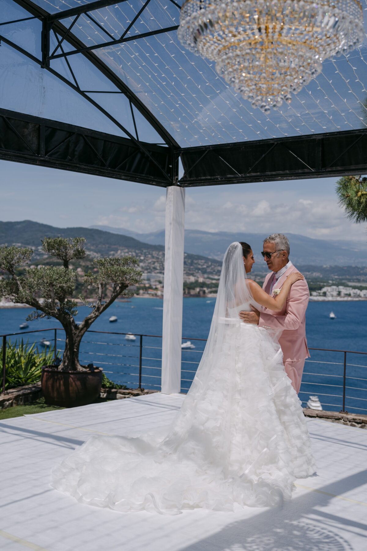 La mariée dans les bras d'un proche, sous une verrière dominant la mer, sa robe étalée sur le sol blanc