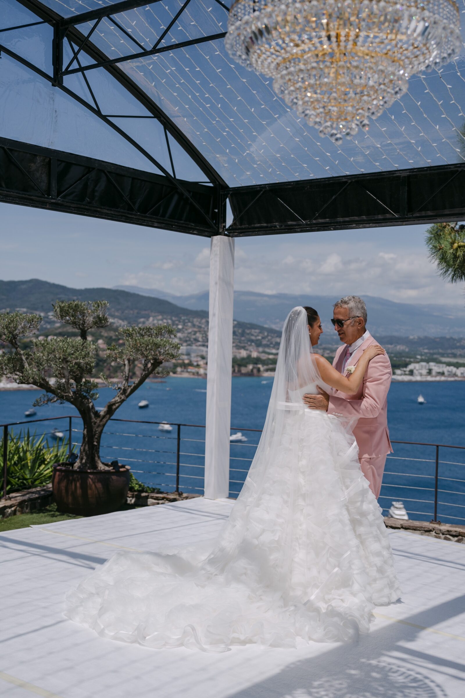 Photographe mariage Saint-Raphaël 4 La mariée dans les bras d'un proche, sous une verrière dominant la mer, sa robe étalée sur le sol blanc