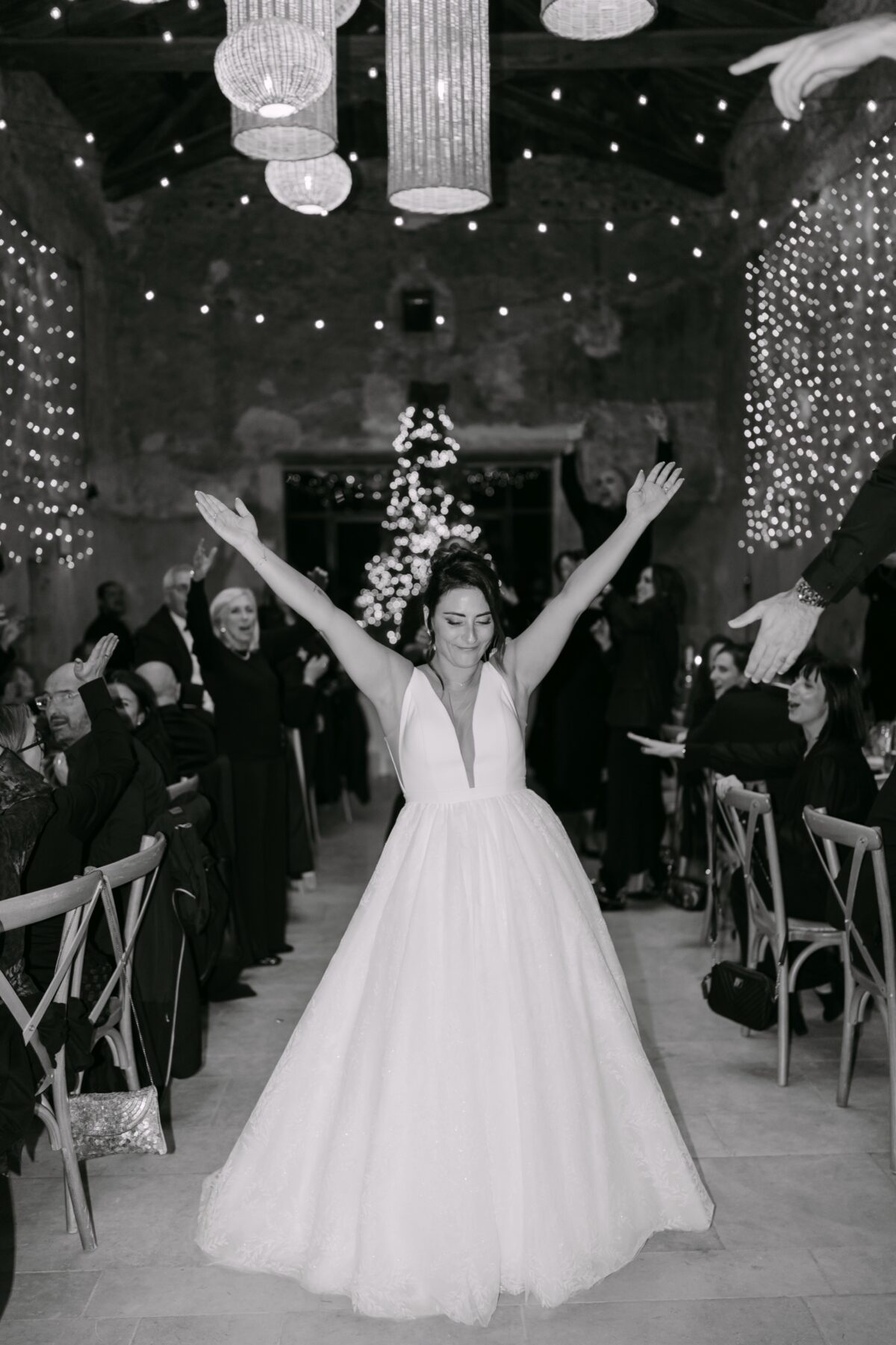En noir et blanc, la mariée lève les bras en entrant dans la salle voûtée, sous les suspensions lumineuses
