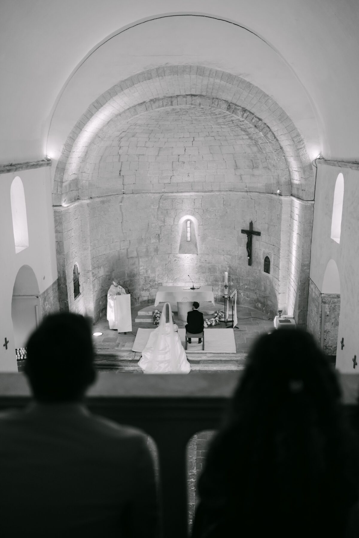 En noir et blanc, la voûte d'une chapelle et les mariés devant l'autel, les invités de dos au premier plan