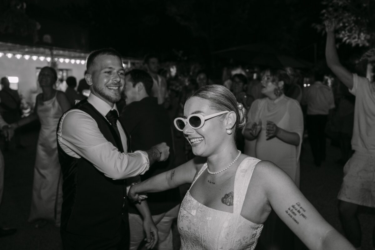 En noir et blanc, la mariée en lunettes blanches danse face au marié au milieu des invités