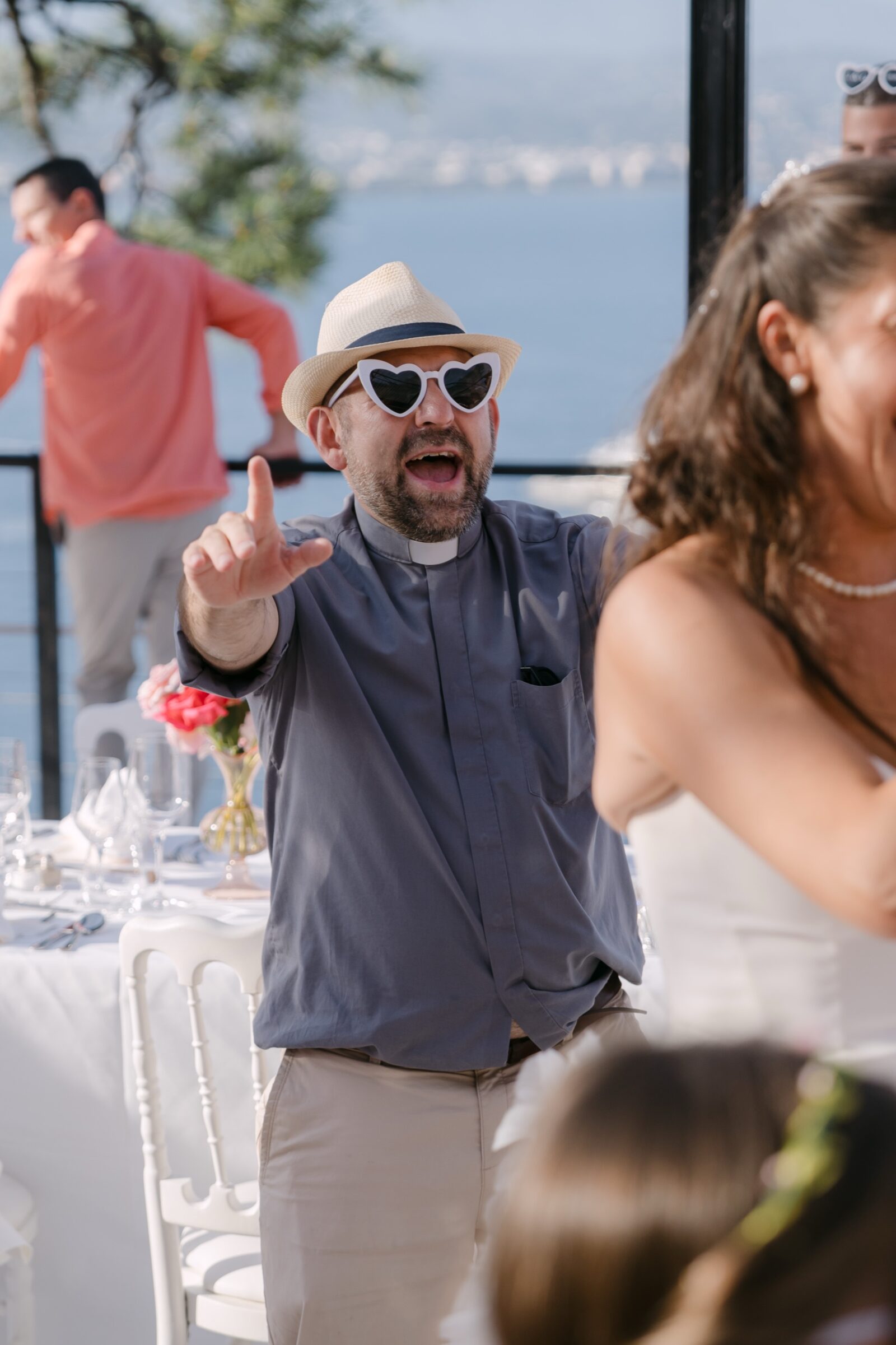 Photographe mariage Côte d’Azur 6 Un invité en chapeau et lunettes en cœur montre du doigt en riant, la mer derrière lui