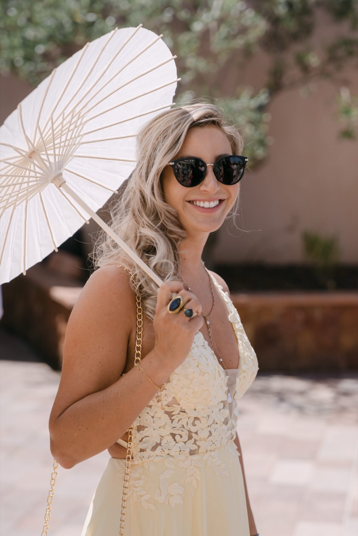 Une invitée en lunettes de soleil sourit sous une ombrelle blanche