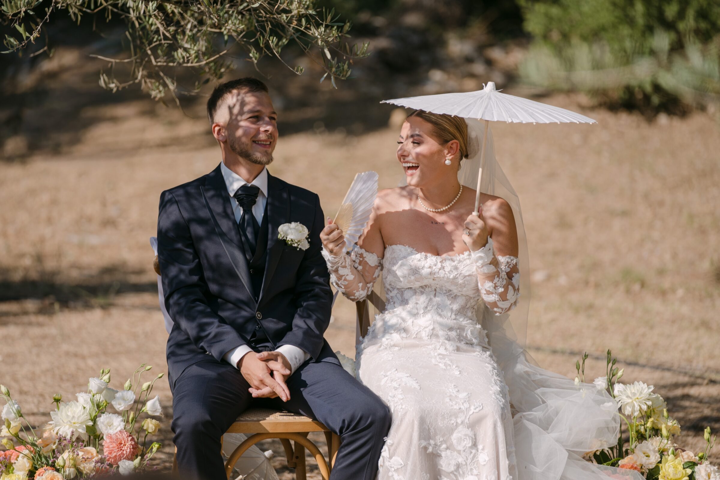 Être naturel sur vos photos de mariage, même si vous détestez ça 4 Pendant la cérémonie, les mariés assis éclatent de rire, la mariée tient une ombrelle