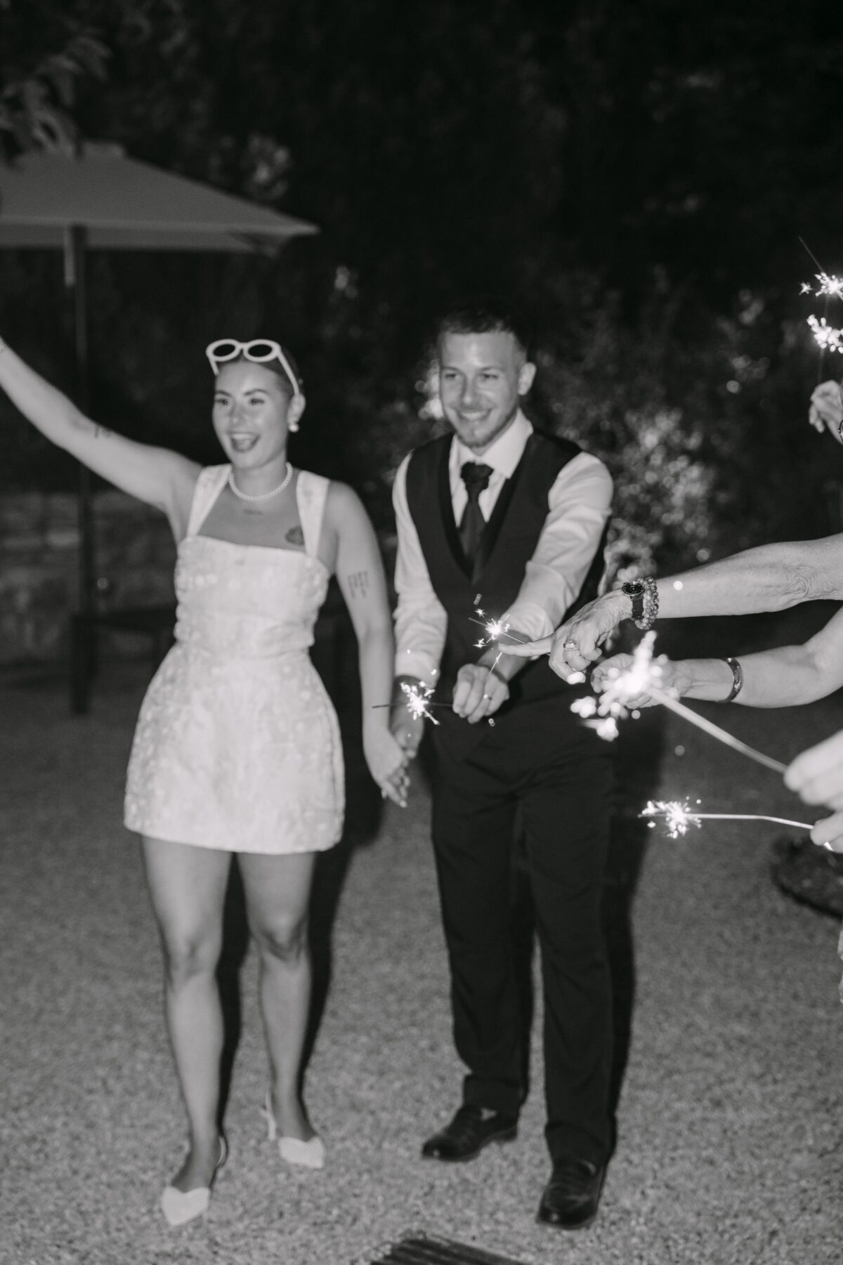 En noir et blanc, les mariés sortent en riant entre les cierges magiques, la mariée en lunettes de soleil